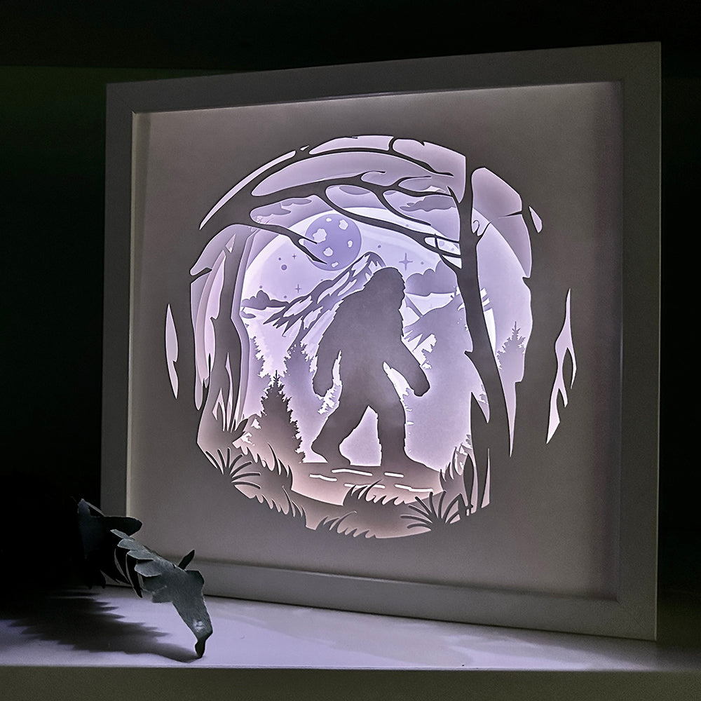Framed paper cutout of a silhouette of a bigfoot in a forest with a moonlit sky, illuminated by a light source.