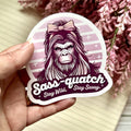Sticker with a bigfoot wearing a bow and text 'Sass-quatch Stay Wild, Stay Sassy' held by a hand with flowers in the background.