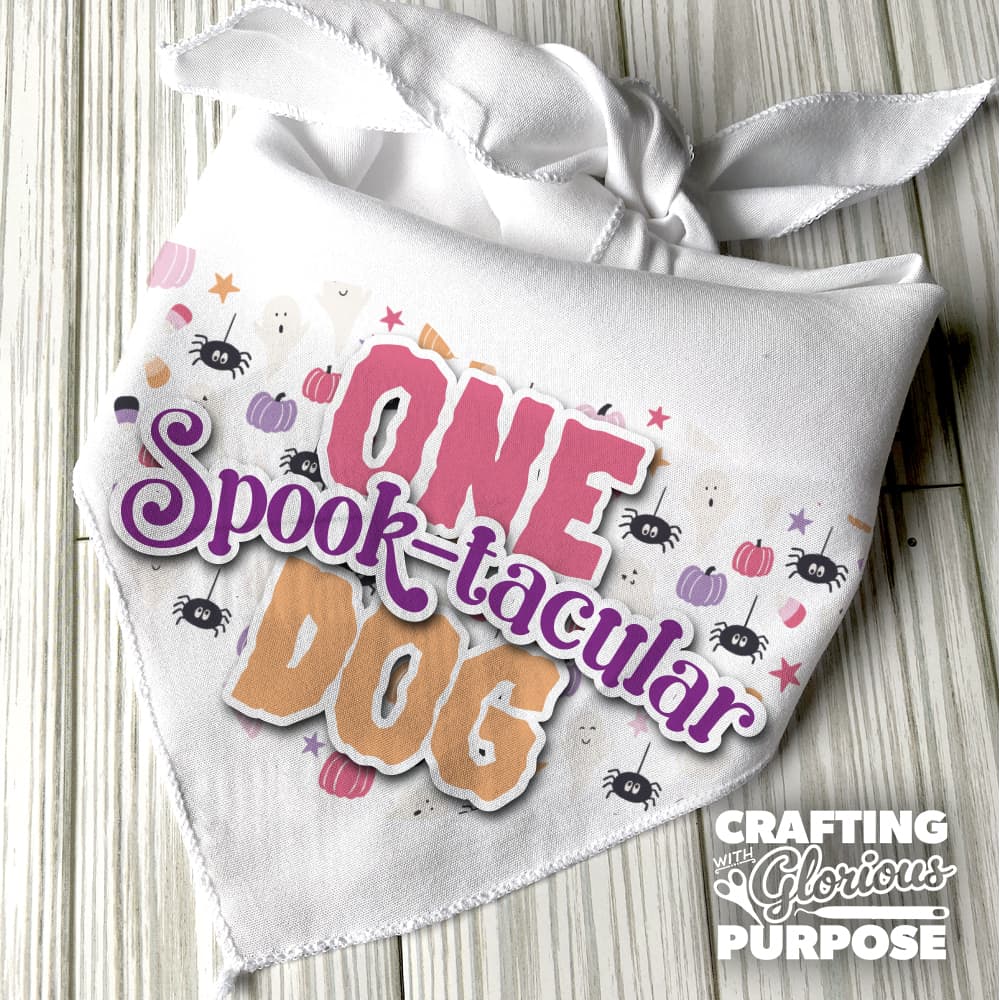 White bandana with 'ONE Spook-tacular Dog' text and Halloween-themed design on a wooden surface.
