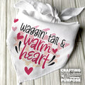 White bandana with text 'Waggin' tail & warm heart' on a wooden surface, featuring the brand 'Crafting with Glorious Purpose'.
