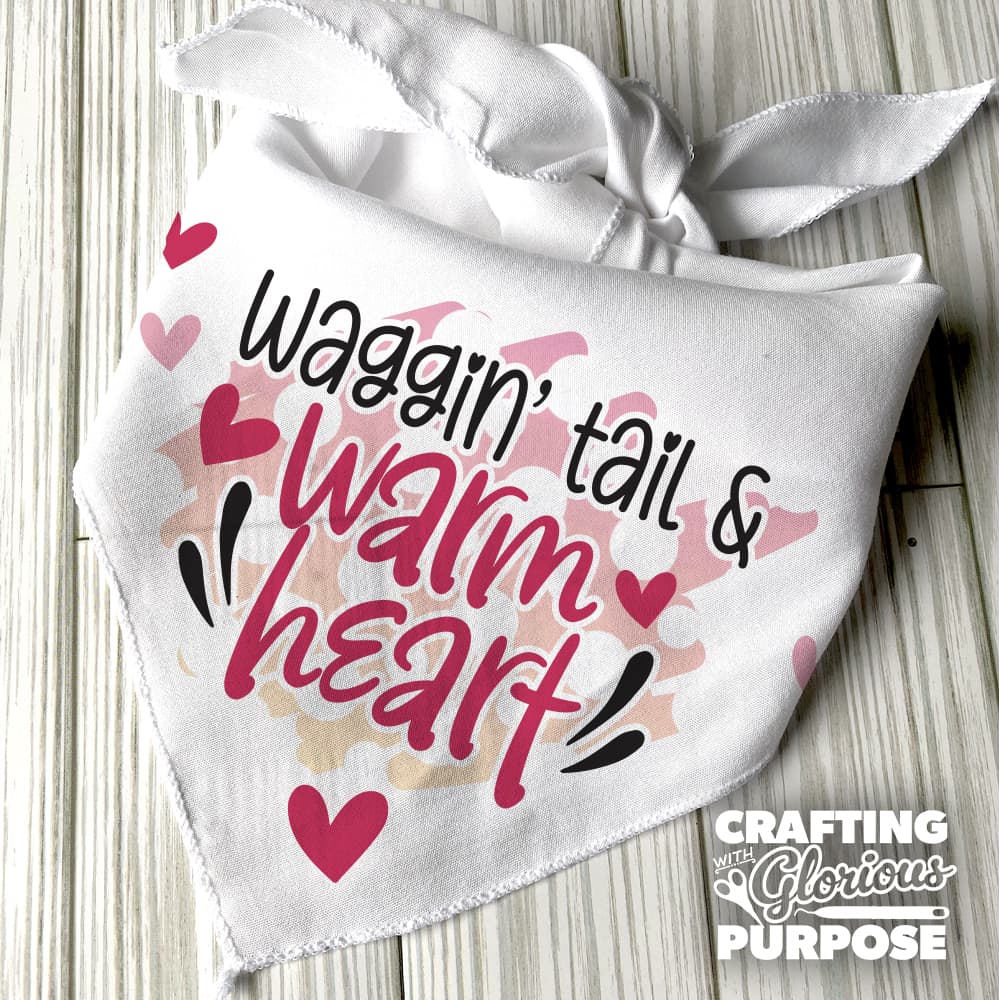 White bandana with text 'Waggin' tail & warm heart' on a wooden surface, featuring the brand 'Crafting with Glorious Purpose'.