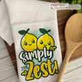 Towel with lemon design and text 'Simply The Zest!' on a wooden surface with a wooden spoon.