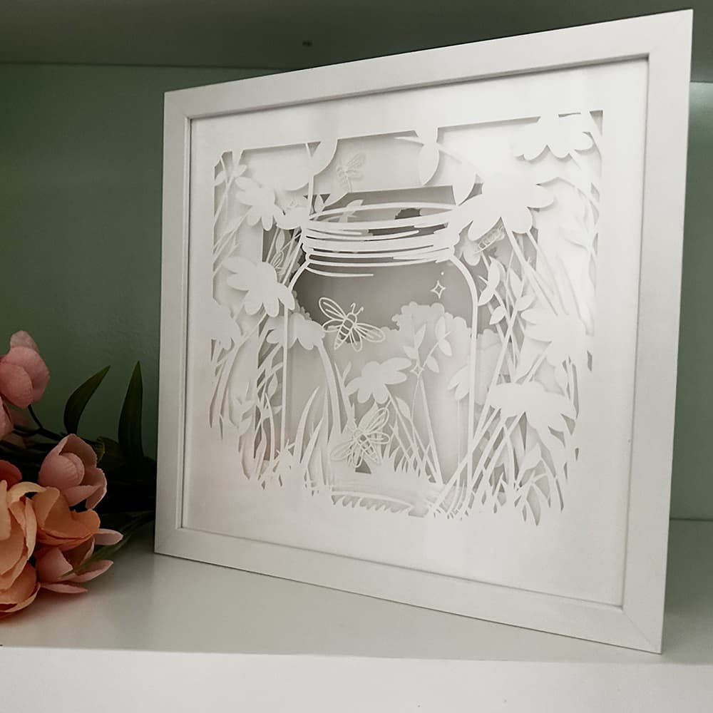 Framed paper cutout art of a jar with flowers and a butterfly on a white surface.