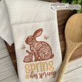 White towel with brown bunny and 'spring has sprung' text on a wooden surface with a wooden spoon.