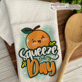 Dish towel with a cartoon peach and 'Squeeze the Day' text on a wooden surface with a wooden spoon.