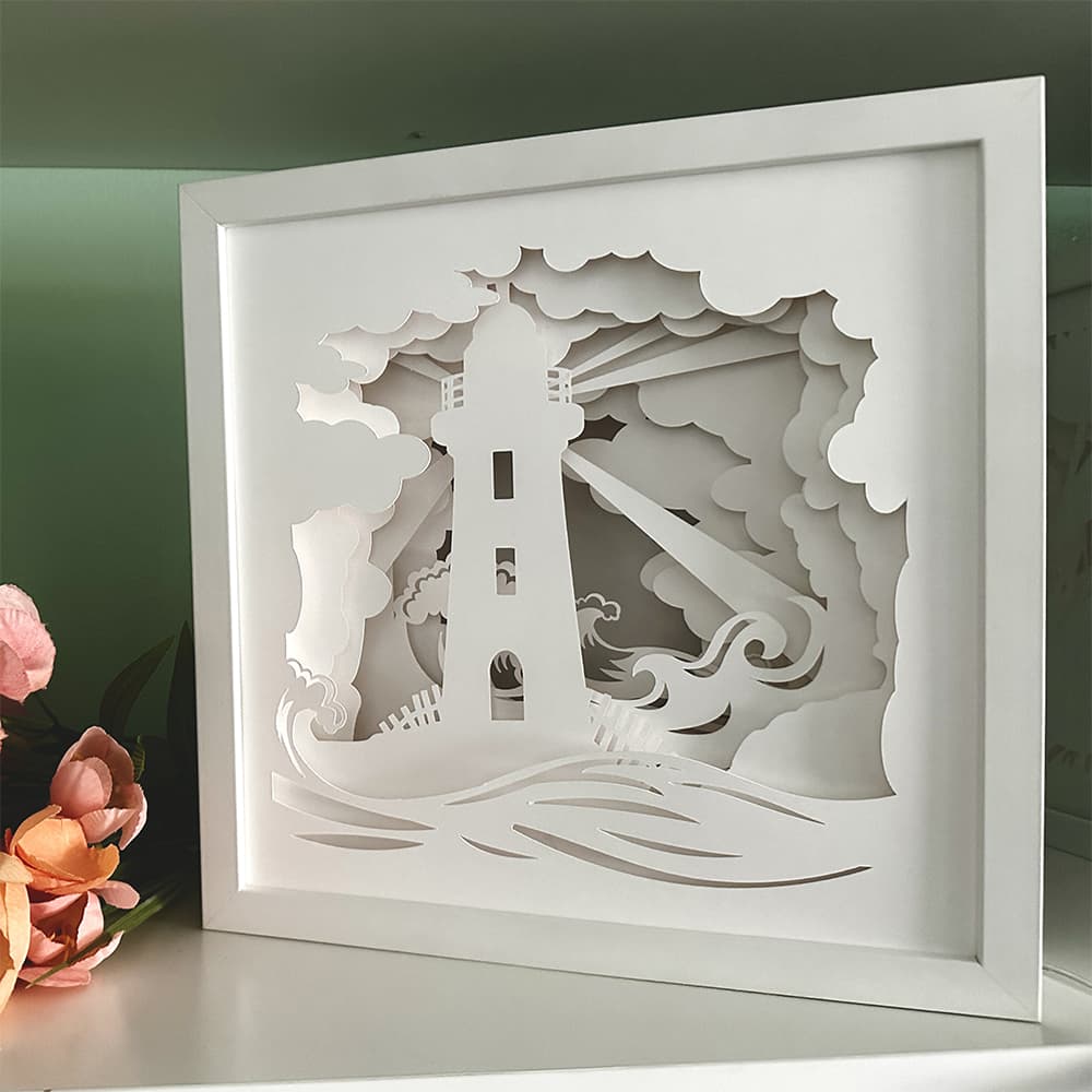 White paper sculpture of a lighthouse with waves and clouds, framed against a green background.