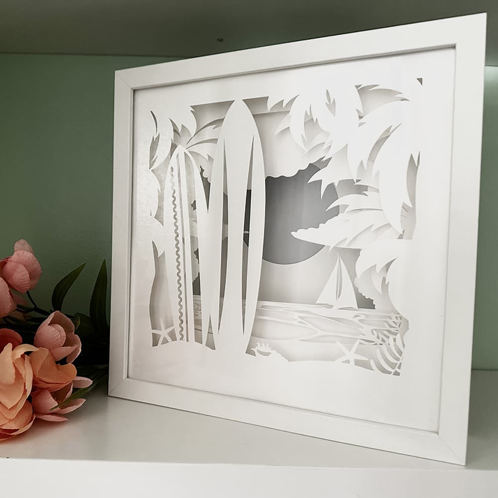 Framed paper cut art of a tropical scene with palm trees and a beach.
