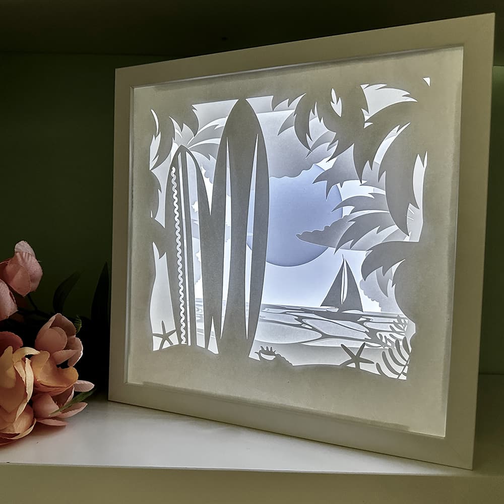 Framed paper cut art of a tropical scene with palm trees and a boat.