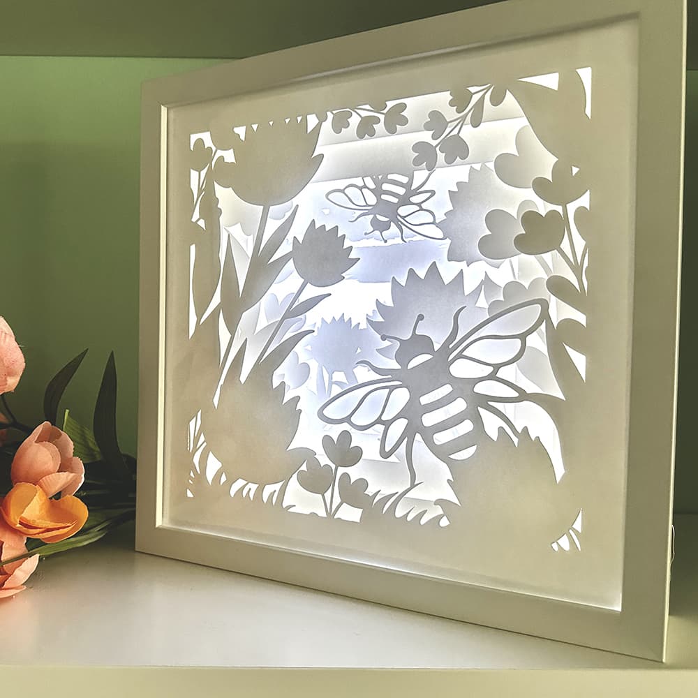 Framed paper art with cut-out designs of bees and flowers on a light surface.