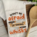 Kitchen towel with 'Don't be afraid of taking whisks' text on a wooden surface with a wooden spoon.