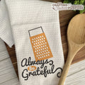 White towel with grater design and 'Always Be Grateful' text on a wooden surface with a wooden spoon.