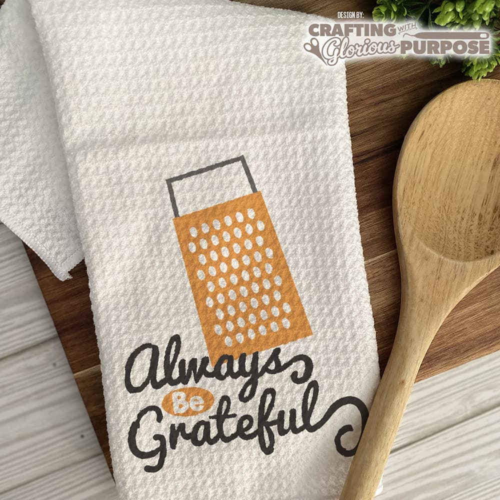 White towel with grater design and 'Always Be Grateful' text on a wooden surface with a wooden spoon.