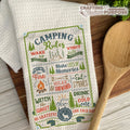Camping-themed towel with text and graphics on a wooden surface with a wooden spoon.