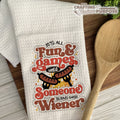 White towel with humorous text design featuring a hot dog, wooden spoon, and text 'It's all fun & games until someone burns their wiener'.