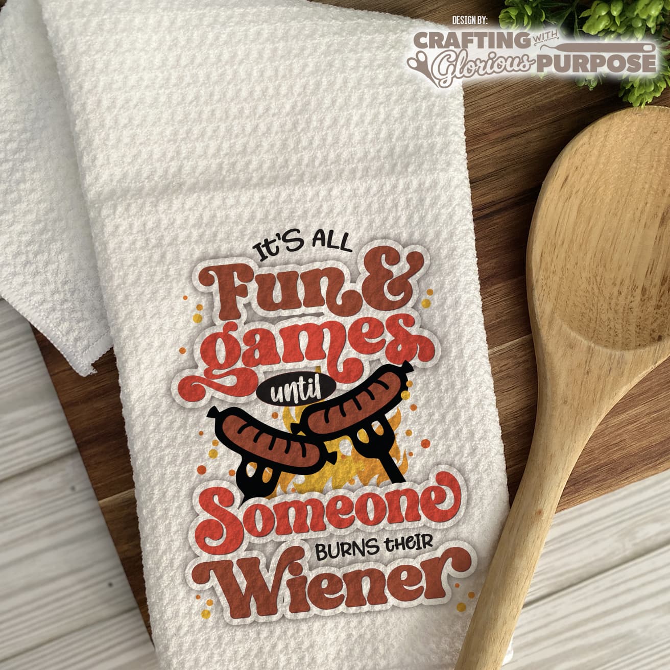 White towel with humorous text design featuring a hot dog, wooden spoon, and text 'It's all fun & games until someone burns their wiener'.