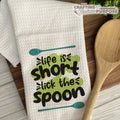White towel with 'life is short, lick the spoon' text and a wooden spoon on a wooden surface.