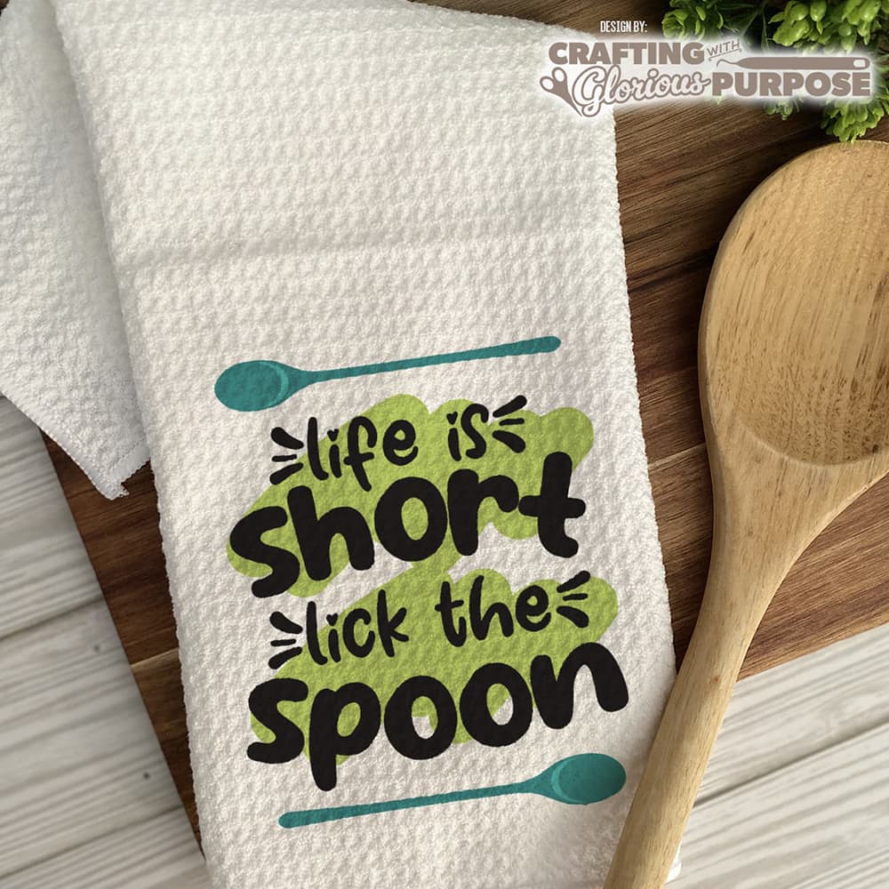 White towel with 'life is short, lick the spoon' text and a wooden spoon on a wooden surface.