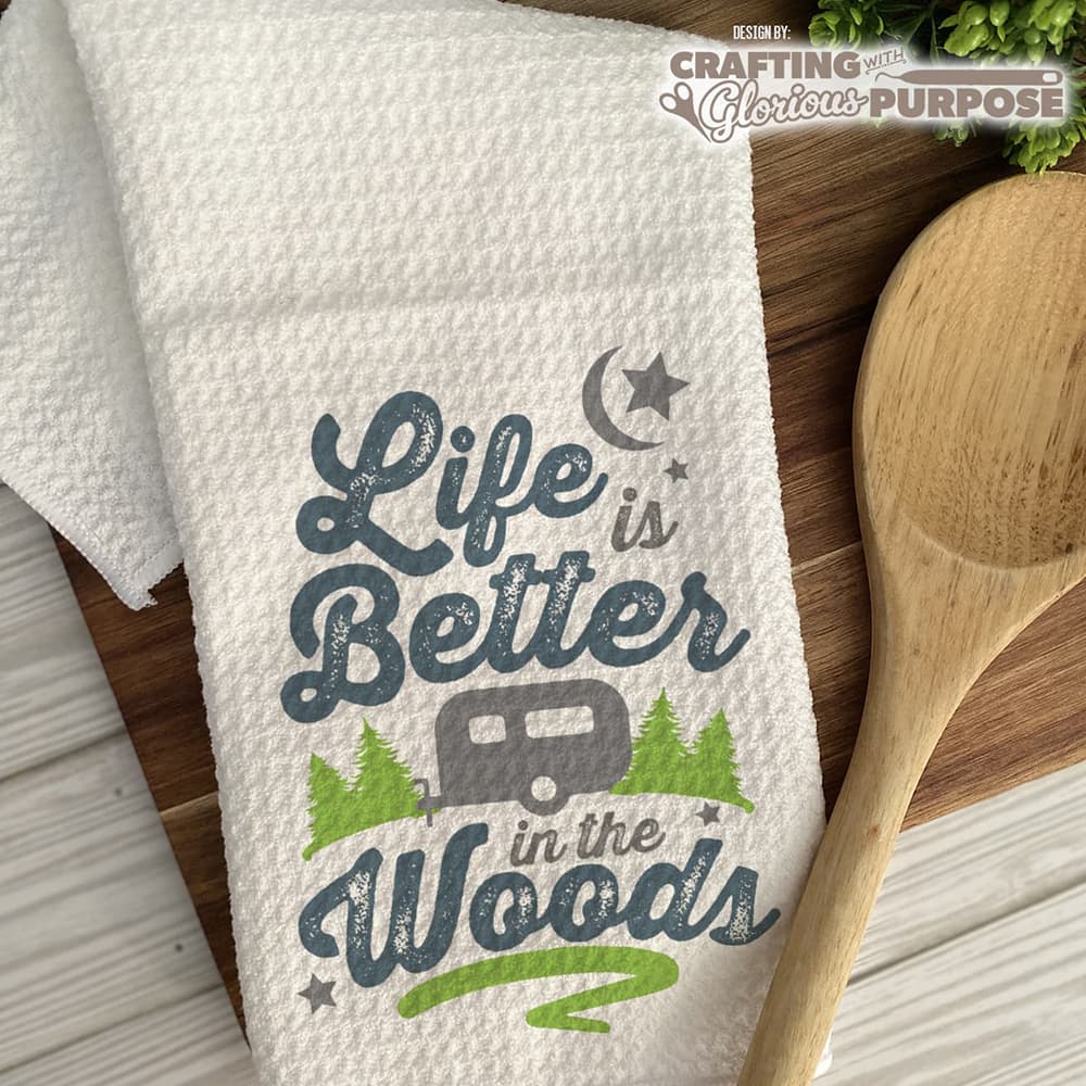 Towel with 'Life is Better in the Woods' text and camper graphic on a wooden surface with a wooden spoon.