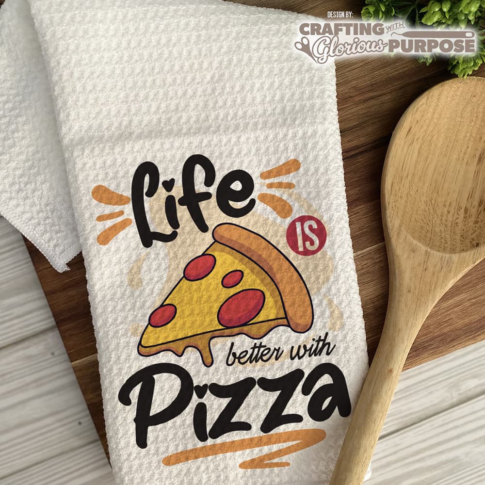 Dish towel with pizza design and text 'Life is better with Pizza' on a wooden surface with a wooden spoon.