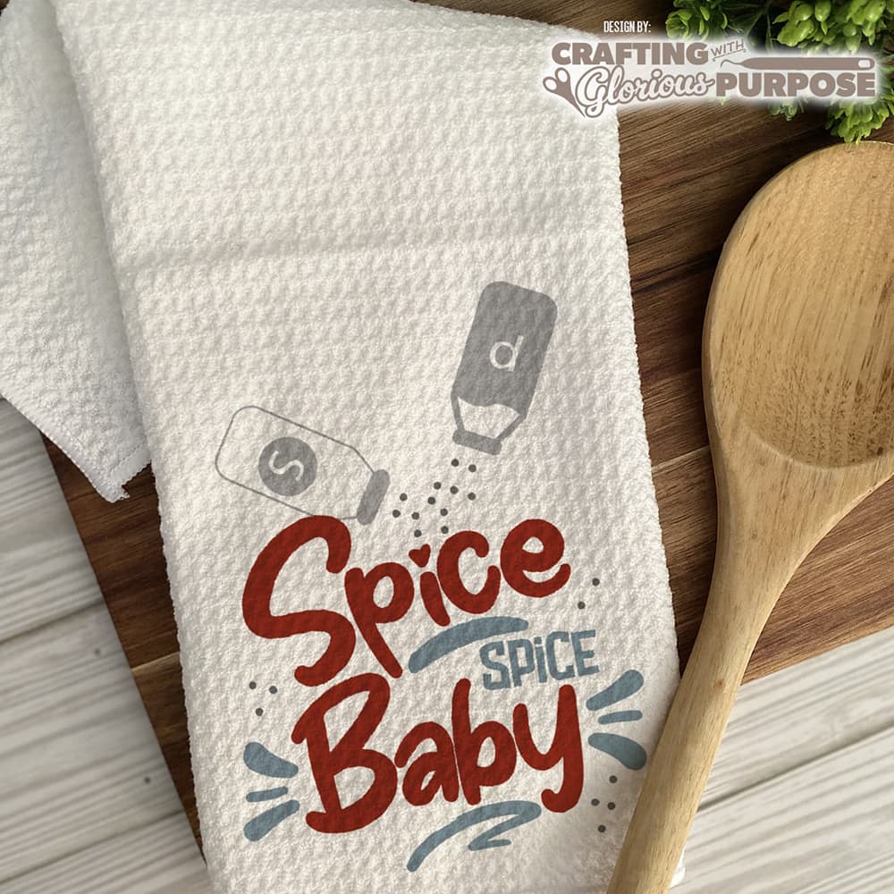 White towel with 'Spice Spice Baby' text and salt and pepper shaker graphics on a wooden surface with a wooden spoon.