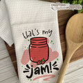 Kitchen towel with a jar of jam design and text, placed on a wooden surface with a wooden spoon.