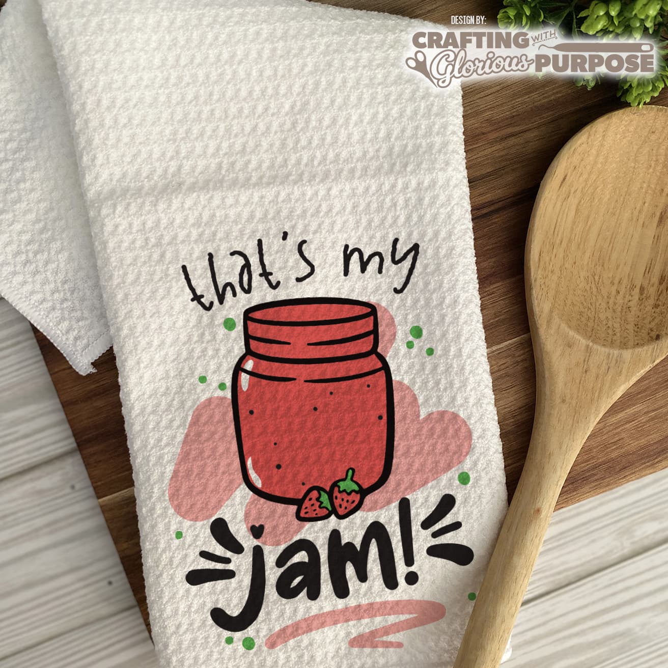 Kitchen towel with a jar of jam design and text, placed on a wooden surface with a wooden spoon.