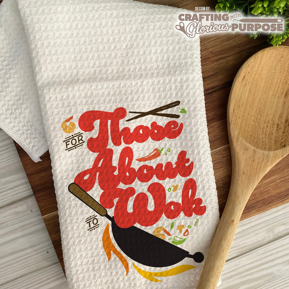 White dish towel with red text and wok illustration on a wooden surface with a wooden spoon.