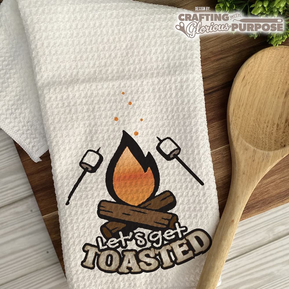 White towel with 'Let's Get Toasted' design on a wooden surface with a wooden spoon.