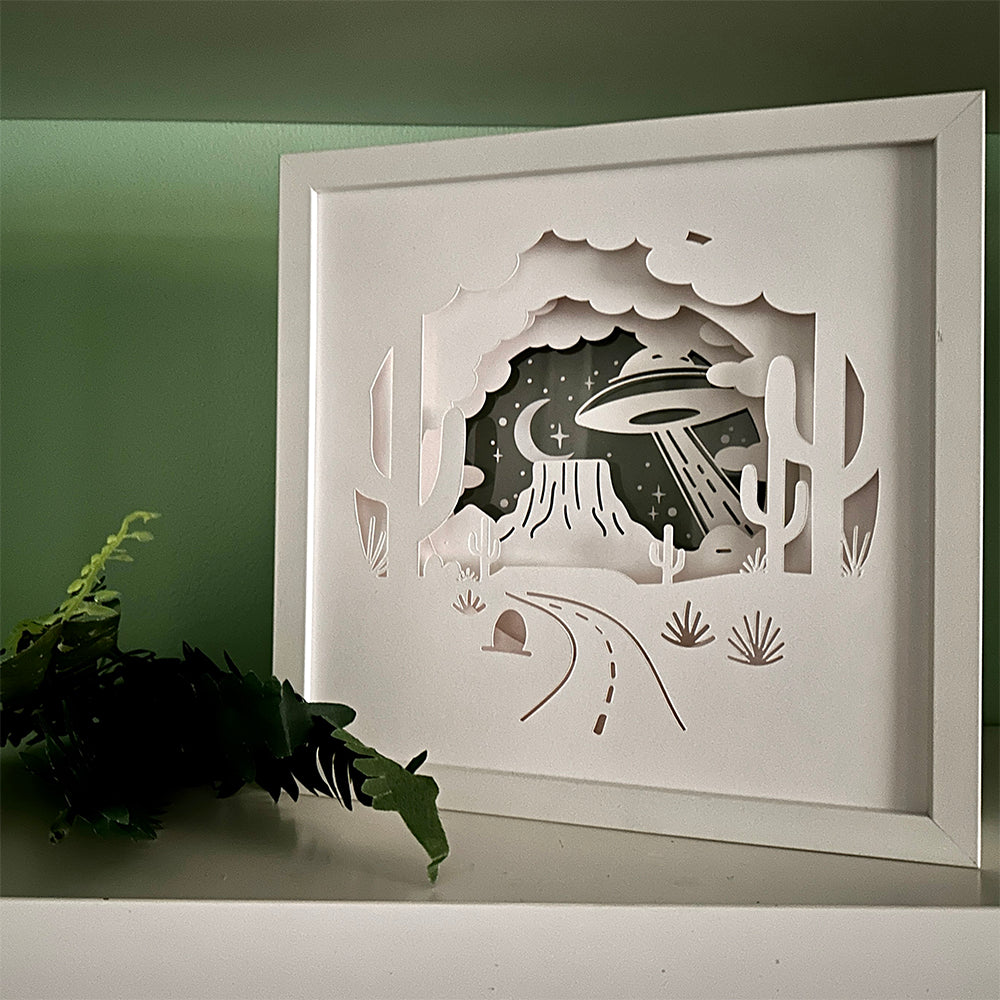 Framed paper cutout art of a UFO over a cactus landscape on a green wall.