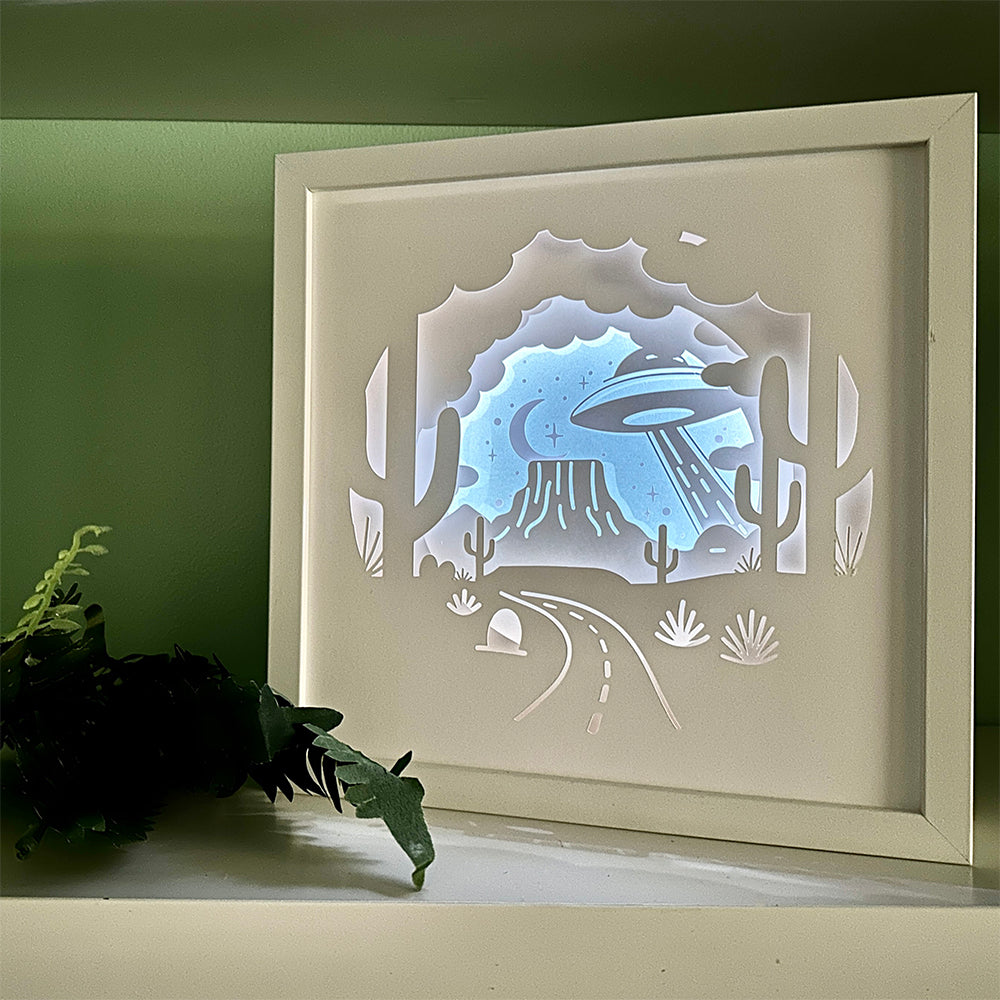Framed paper cutout art of a UFO over a desert landscape with a green wall background.