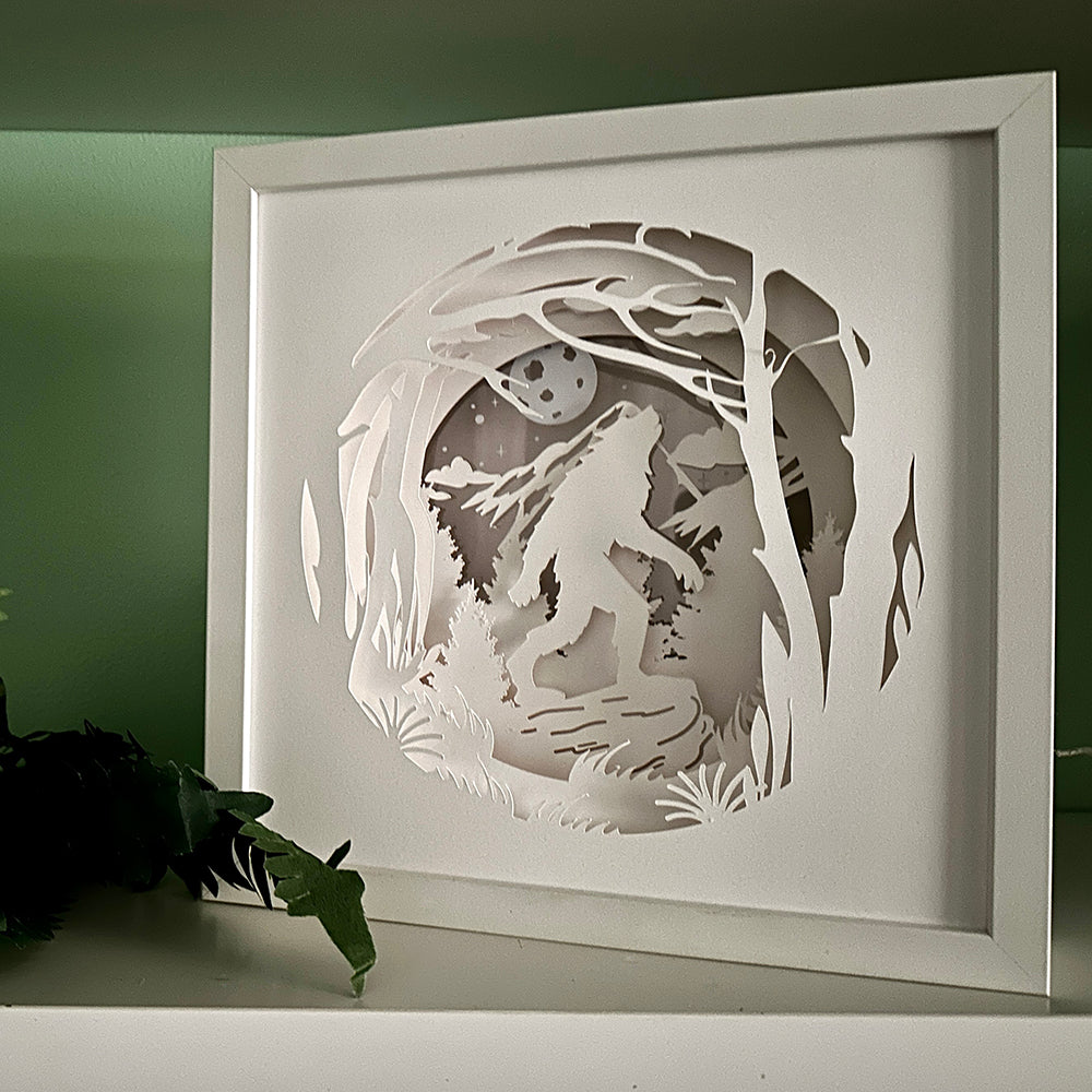 Framed paper cutout art of a wolf in a forest with a moonlit sky.