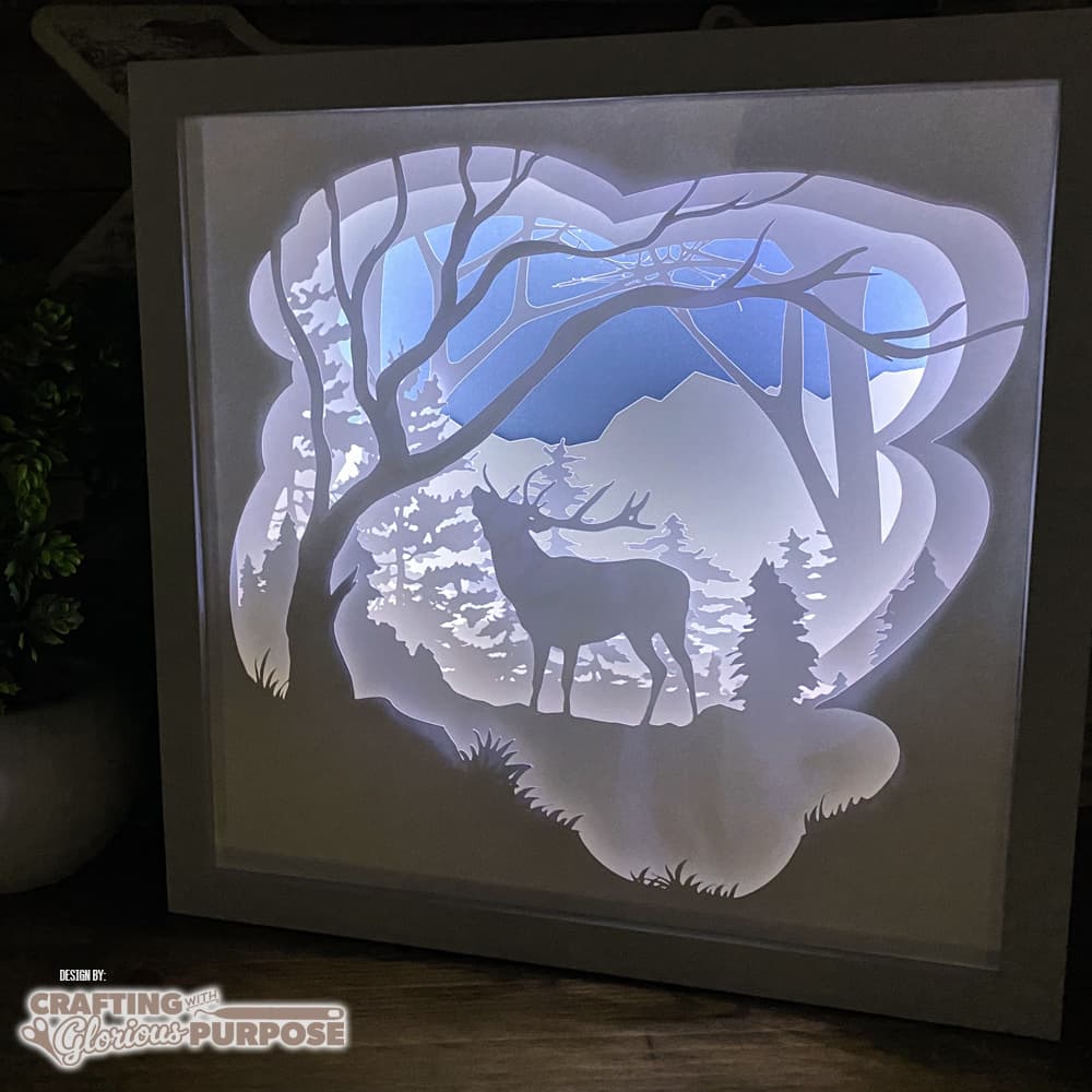 Framed paper cut art of a deer in a forest scene with lighting.