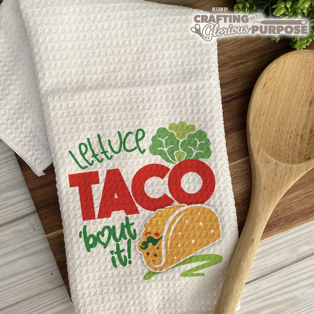 White towel with taco design and text on a wooden surface with a wooden spoon.