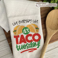 White towel with 'Live everyday like it's Taco Tuesday!' text and taco graphics on a wooden surface with a wooden spoon.