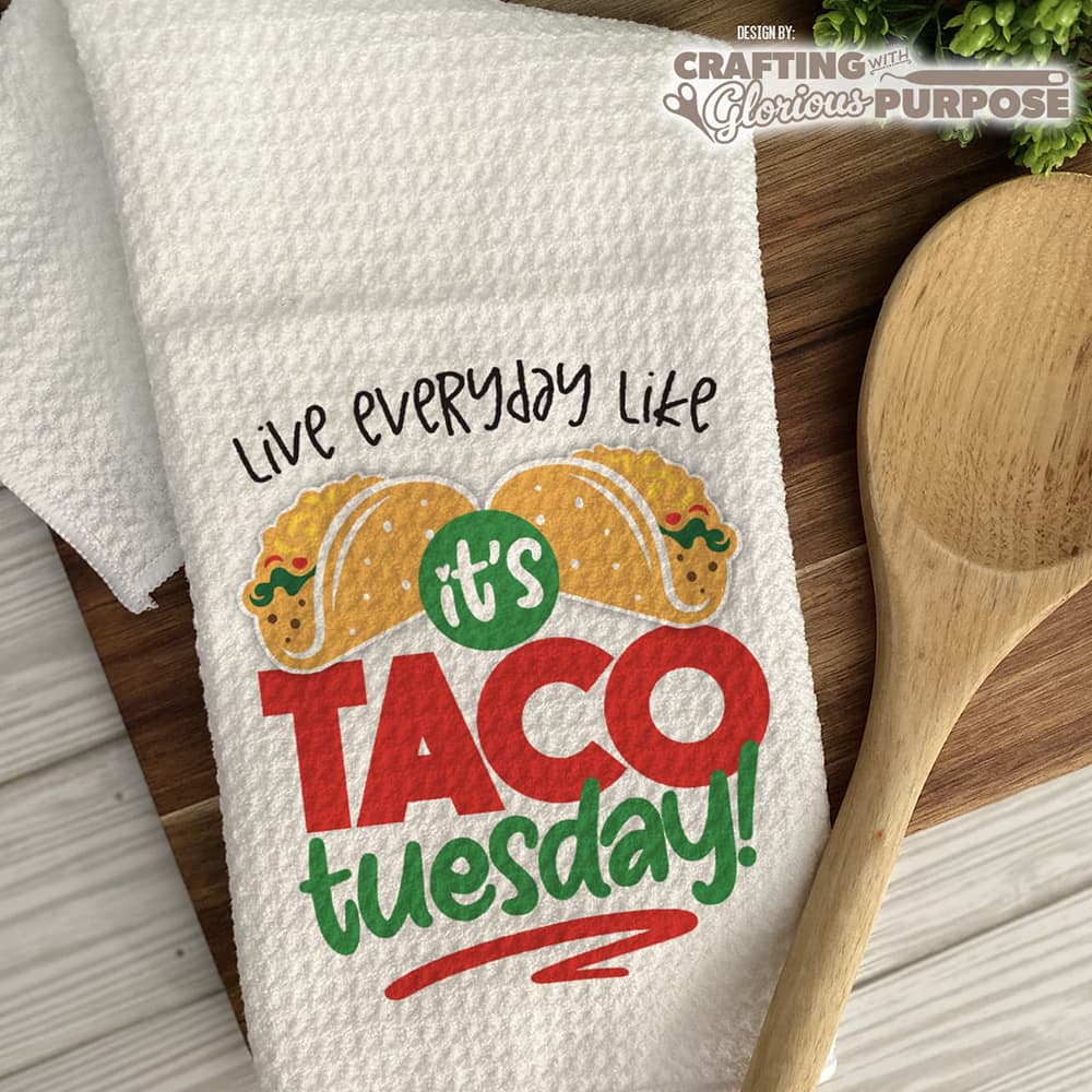 White towel with 'Live everyday like it's Taco Tuesday!' text and taco graphics on a wooden surface with a wooden spoon.